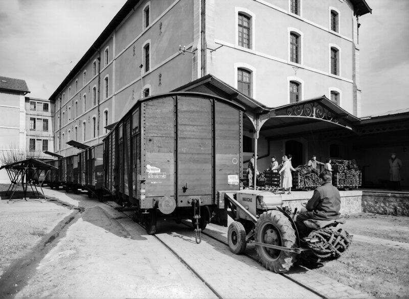 Des ouvrières chargent des caisses de tabac depuis un quai couvert vers une rame de wagons de bois ; un tracteur de manœuvre en bout de voie.